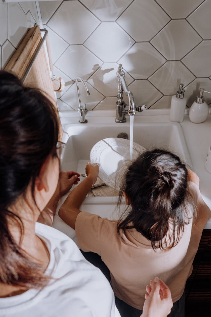 A mother teaching her child dishwashing duties at the kitchen sink.