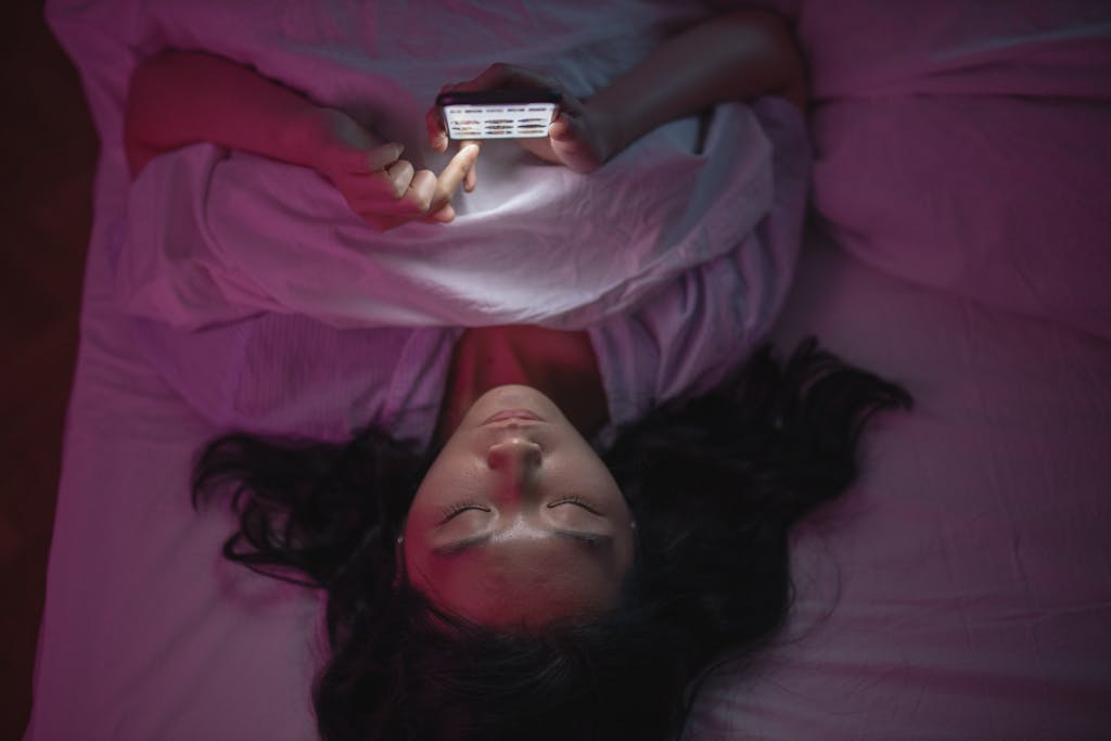 A woman lying in bed at night browsing her smartphone under soft lighting.