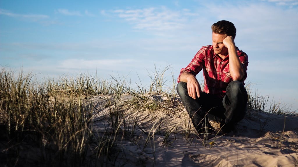 Man in a red plaid shirt sitting thoughtfully on a sandy dune in Australia, surrounded by grass.